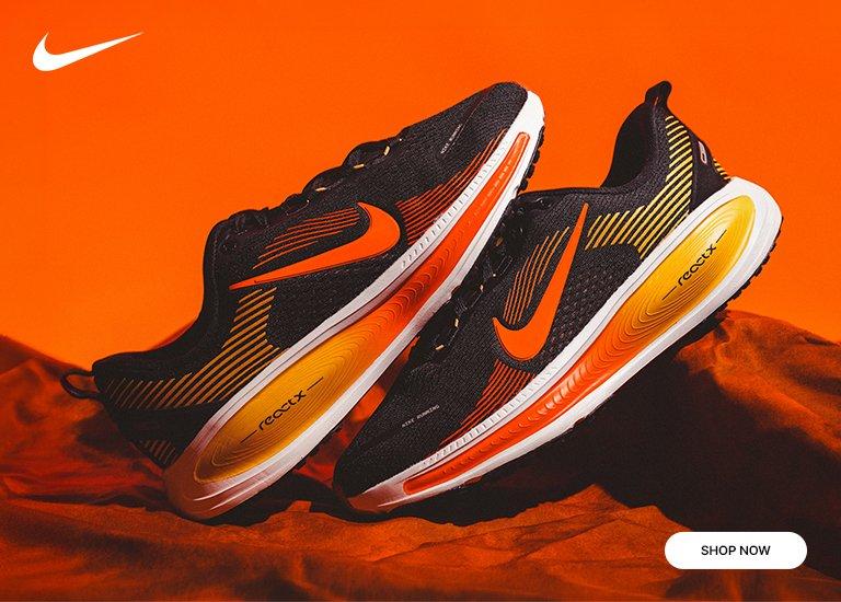 Promotional banner featuring two black Nike running sneakers with vibrant orange swooshes and yellow-to-white gradient midsoles. The shoes are resting on a draped, orange fabric background. A white Nike logo is in the top-left corner, and a Shop Now button is in the bottom-right.
