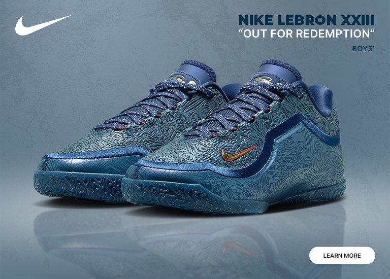 A pair of blue Nike LeBron XXIII Out For Redemption boys' sneakers with intricate patterns and gold accents. The shoes are set against a smoky blue background featuring a white Nike swoosh and a "LEARN MORE" button.