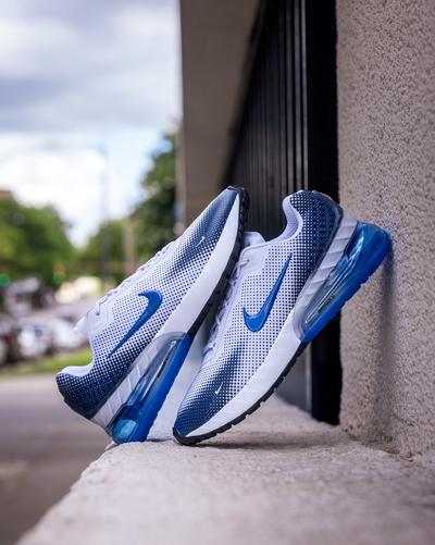  A pair of white and blue Nike sneakers are shown up close, leaning against a textured outdoor wall. The shoes feature a dotted blue gradient pattern on the upper, a bright blue swoosh, and a chunky, translucent blue sole. The background is a blurry street scene.