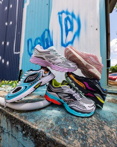An aerial shot of two Saucony sneakers on a two-toned textured ground, split diagonally into pink and green. One sneaker is all pink with white laces, and the other is black with iridescent pink, gold, and blue accents, along with red laces.