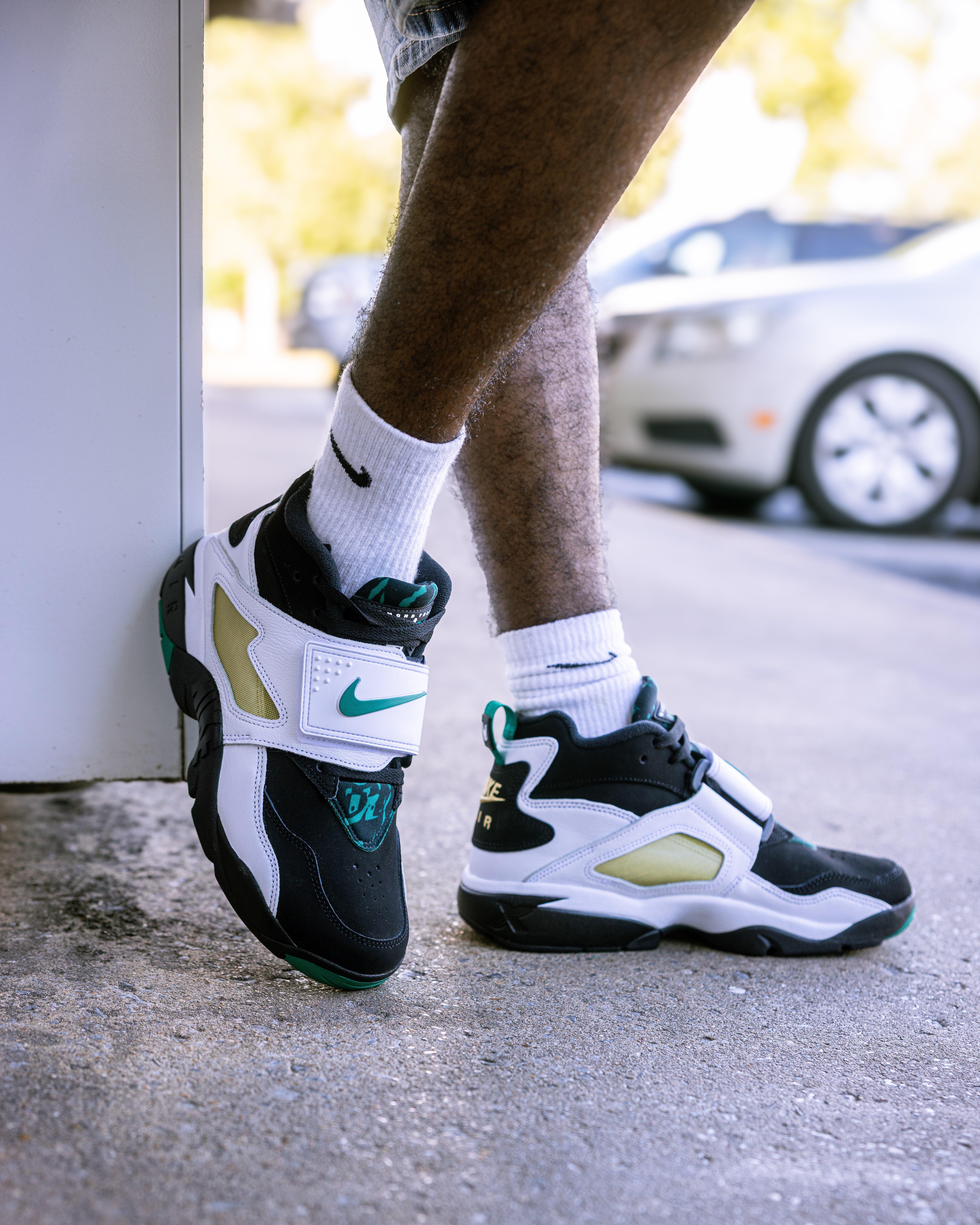 Low-angle, on-foot view of a person wearing Nike Air Diamond Turf sneakers in a black, white, and emerald green colorway. The person is wearing white Nike socks and leaning against a white wall outdoors.