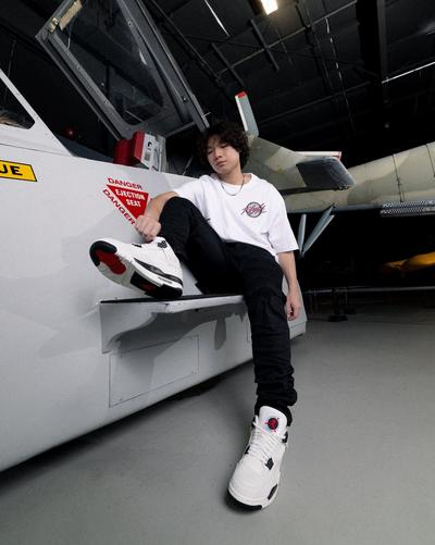 A young man with curly hair sits on the edge of a white aircraft cockpit, wearing a white Jordan Brand t-shirt, black pants, and white Air Jordan 4 sneakers.