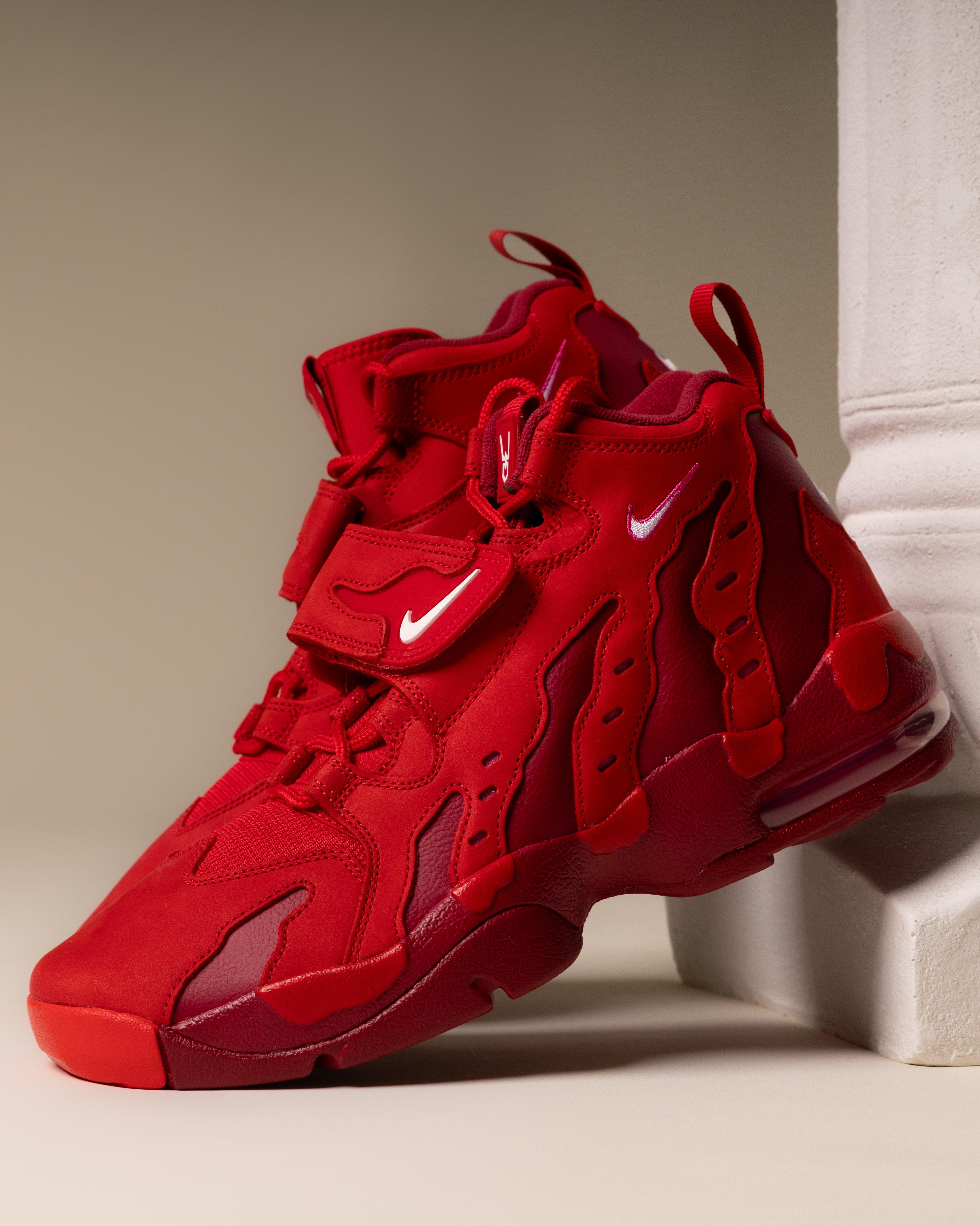 A pair of all-red Nike Air DT Max '96 sneakers leaning against a white classical-style pillar. The shoes feature a prominent midfoot strap with a white Swoosh and signature wavy "shred" overlays.