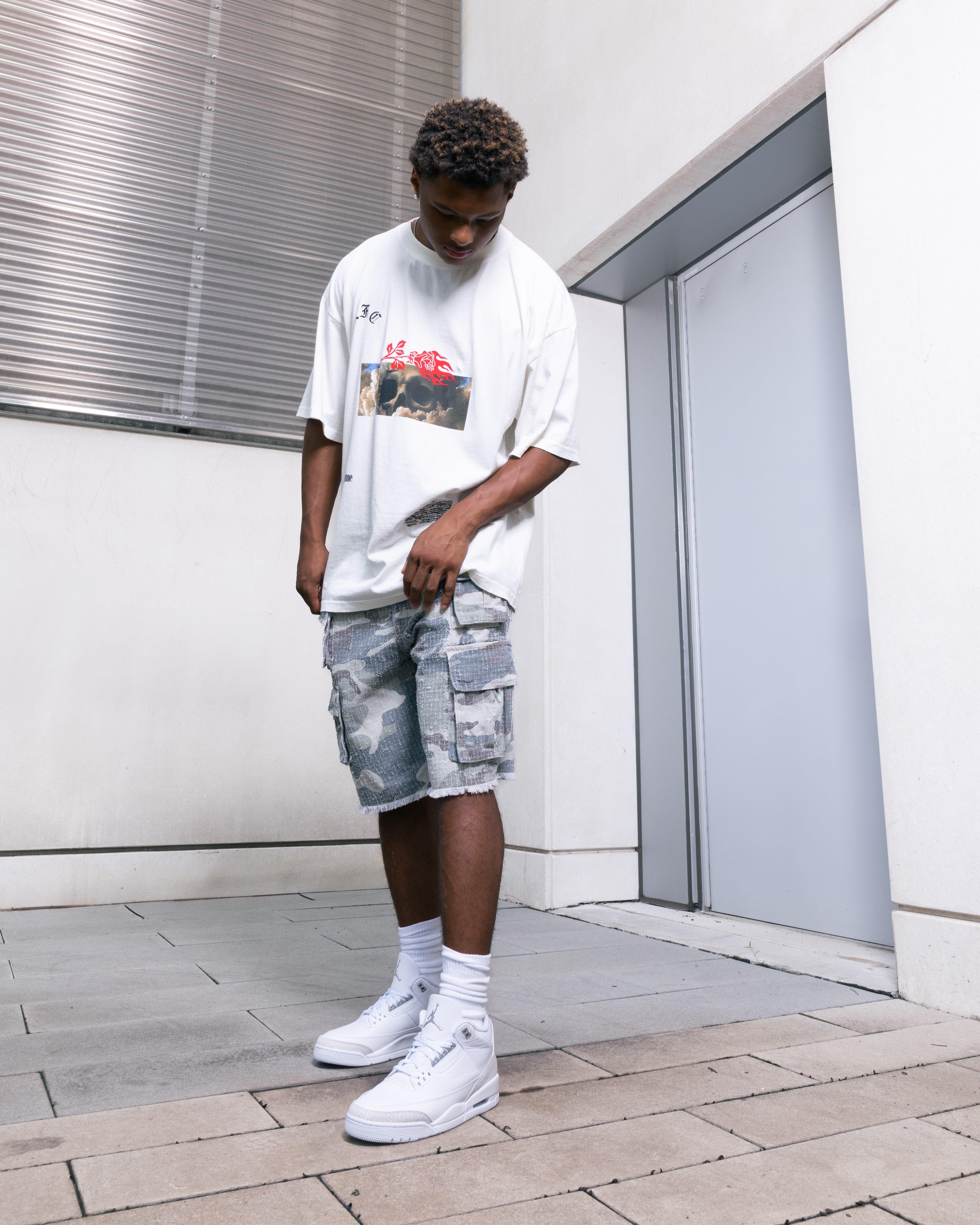 A person's lower legs and feet, wearing white high-top sneakers, white crew socks, and light-colored distressed camouflage shorts. The sneakers are predominantly white with subtle grey textured accents, and a silver Jumpman logo is visible on the tongue."