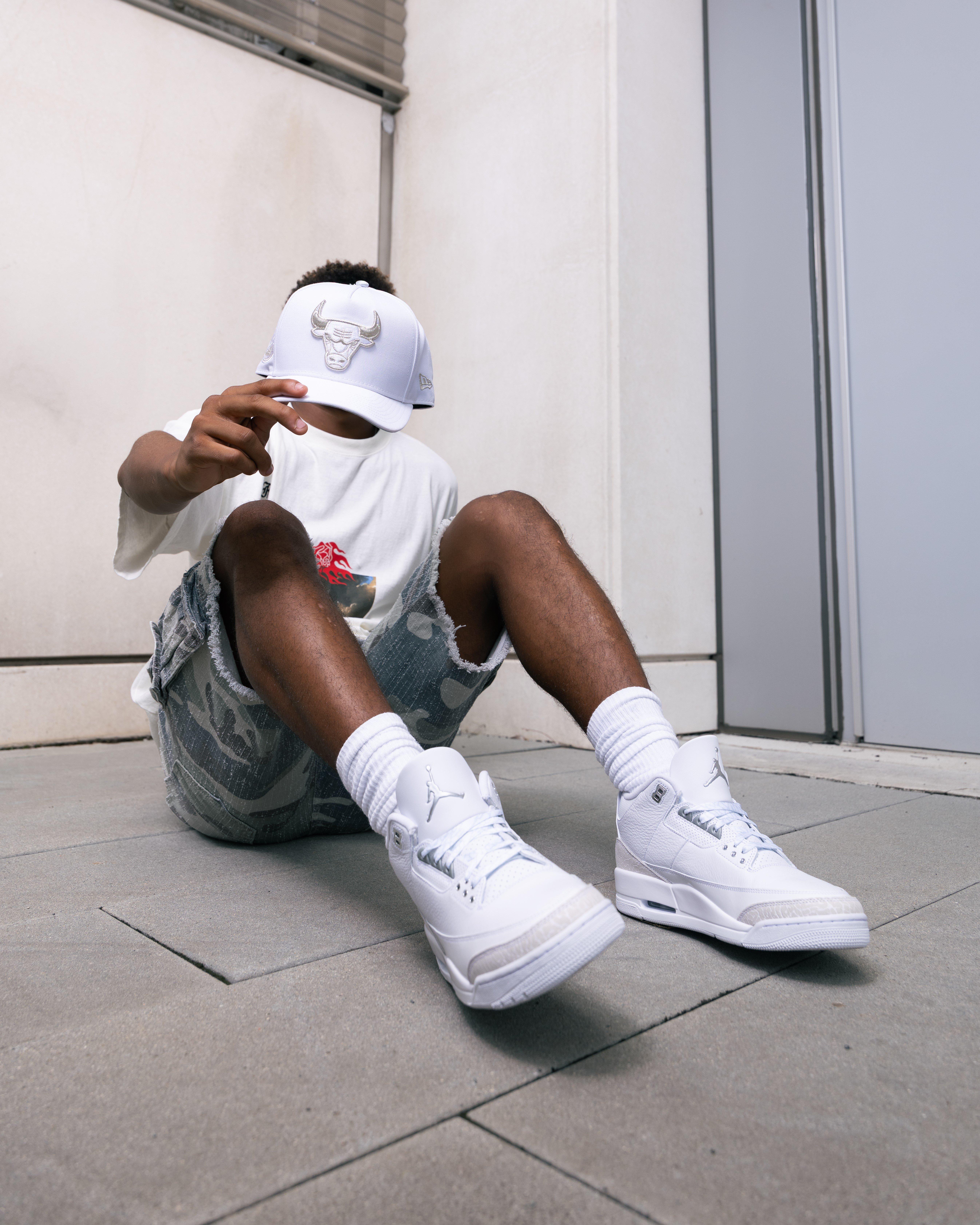 A person is sitting on a concrete floor, leaning against a light-colored wall. They are wearing white high-top sneakers, white crew socks, distressed light-colored shorts, a white t-shirt, and a white baseball cap with a Chicago Bulls logo. Their face is obscured by the cap.