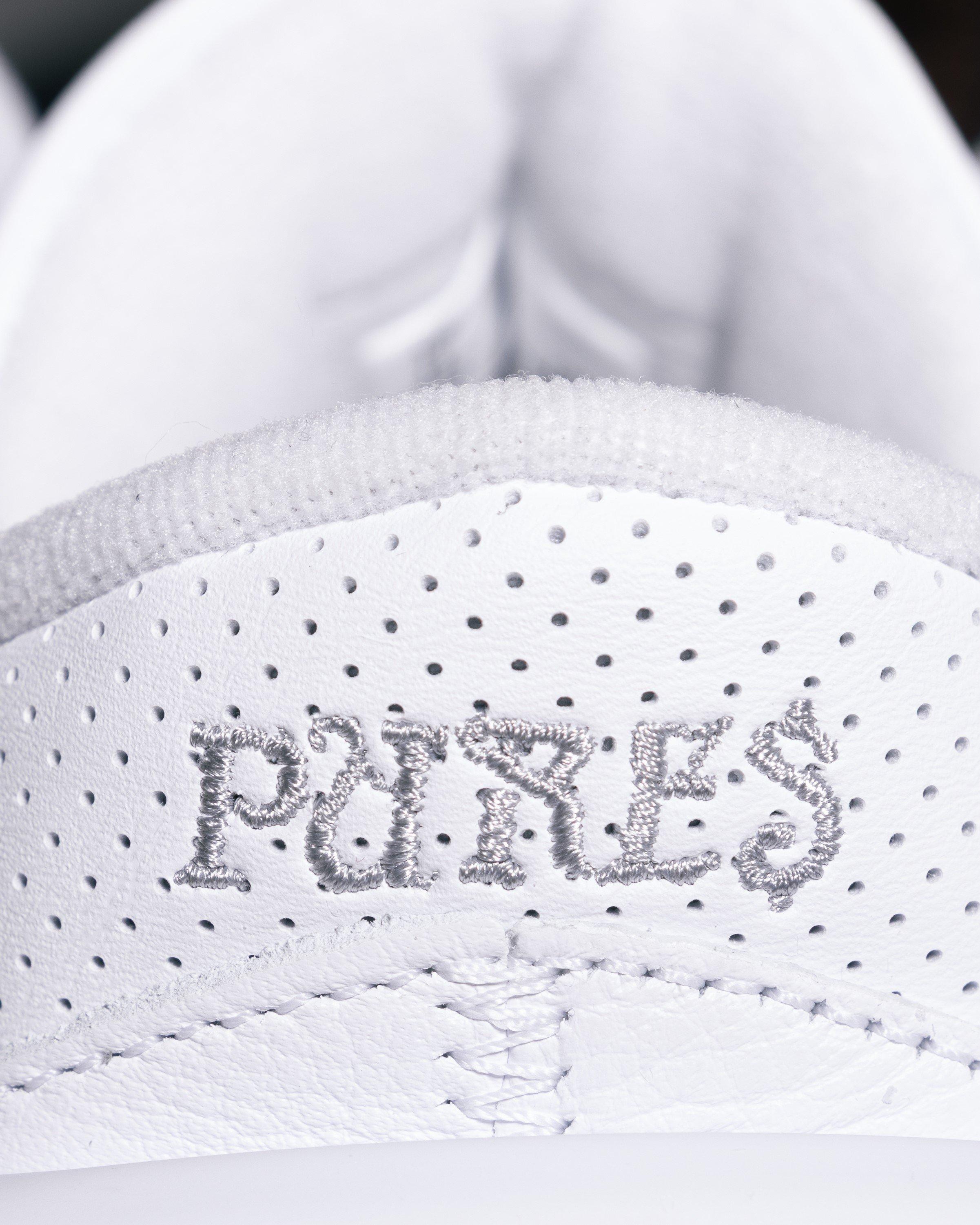 A close-up shot of the heel collar of a white sneaker. The word 'PURE$' is embroidered in silver, textured lettering on a perforated white leather or synthetic material. The top edge of the collar is also textured white.