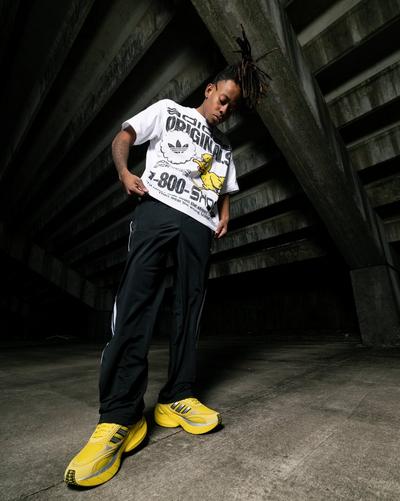 A man with dreadlocks stands in a concrete parking garage wearing a white graphic Adidas Originals t-shirt, black track pants, and chunky yellow Adidas sneakers. Low-angle shot focusing on the vibrant footwear against an industrial background.