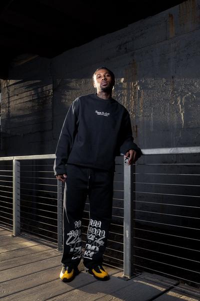 Man wearing a black sweatshirt and joggers with yellow sneakers, leaning against a railing in an industrial urban setting.