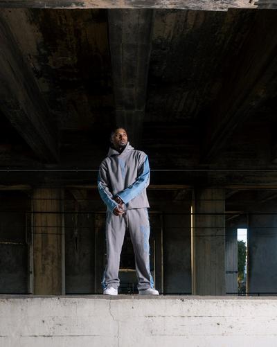 A man stands under a dark concrete overpass wearing a grey hooded tracksuit with blue denim patchwork details on the sleeves and legs. The outfit features light blue embroidered text. He is looking off to the side, standing on a concrete ledge against a background of massive support pillars.