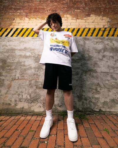 A person with curly dark hair standing against a weathered brick and concrete wall, looking downward with one hand behind their head. They are wearing a white graphic t-shirt featuring a yellow race car and the text "US AGAINST TIME," black shorts, white mid-calf socks, and white sneakers. The background features a yellow and black hazard-striped line on the wall and a red brick ground.