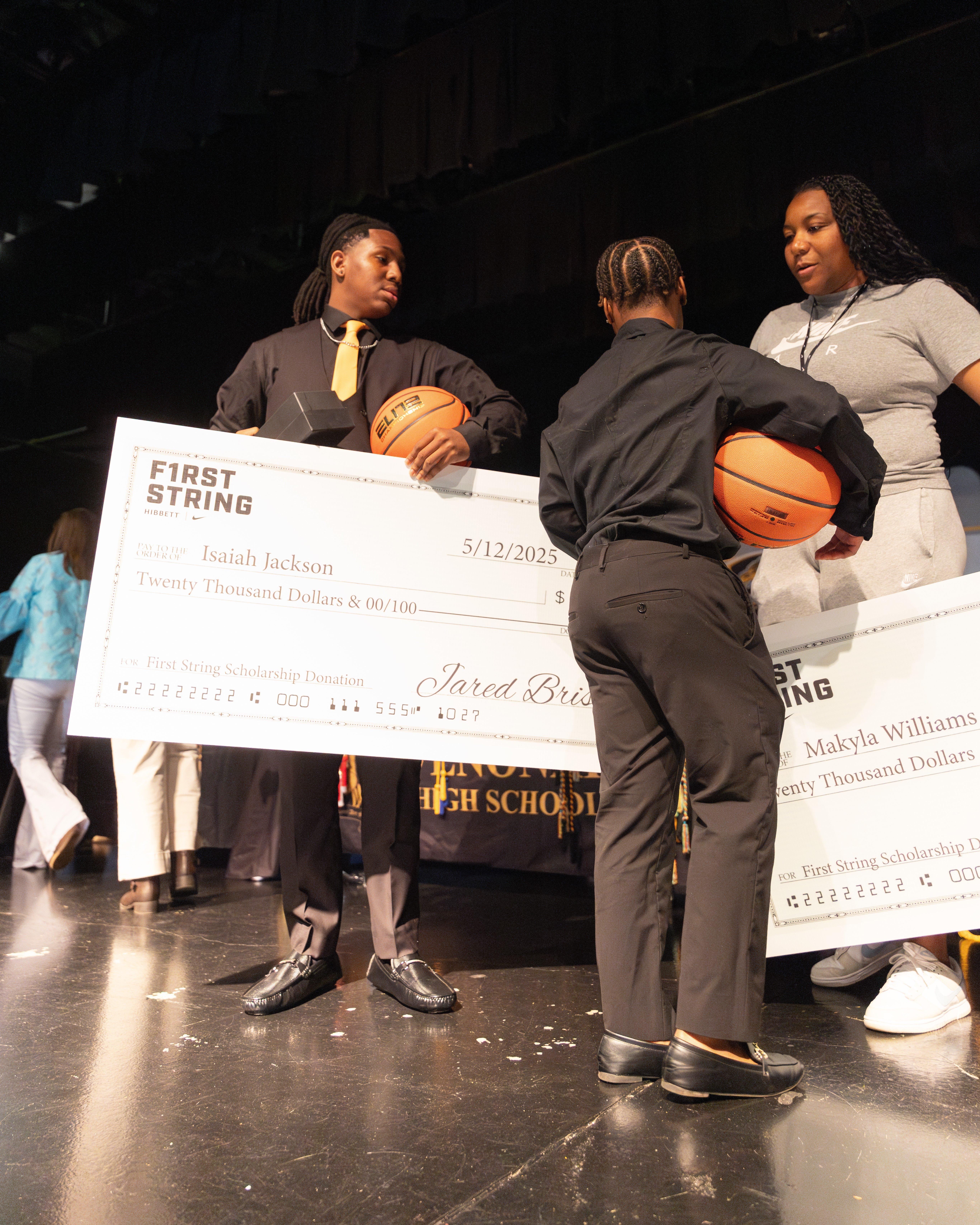 Two young students, dressed in button-up shirts and ties, stand on a stage holding oversized scholarship checks from 'First String Scholarship Donation'. Each student also holds a basketball. Behind them, a woman in a grey t-shirt looks on. The checks are dated '5/12/2025' and are for 'Twenty Thousand Dollars'.