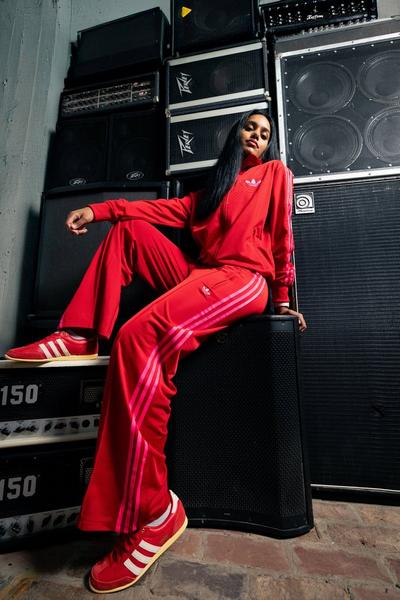 A woman with long dark hair sits on top of a large black speaker, surrounded by a stack of amplifiers and speakers. She is wearing a red Adidas tracksuit with lighter red stripes and matching red sneakers with white stripes.