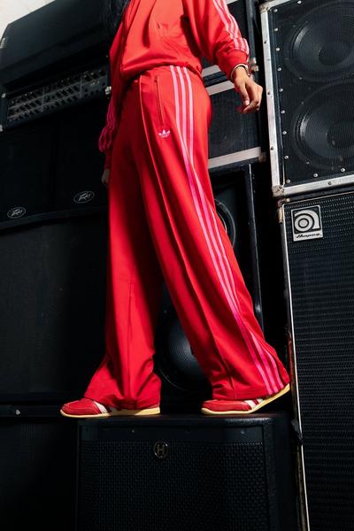 A close-up shot of a person's legs and feet. The person is wearing red Adidas track pants with lighter red stripes, standing on a large black speaker. The speaker is part of a tall stack of audio equipment, and the person is wearing red Adidas sneakers with white stripes.