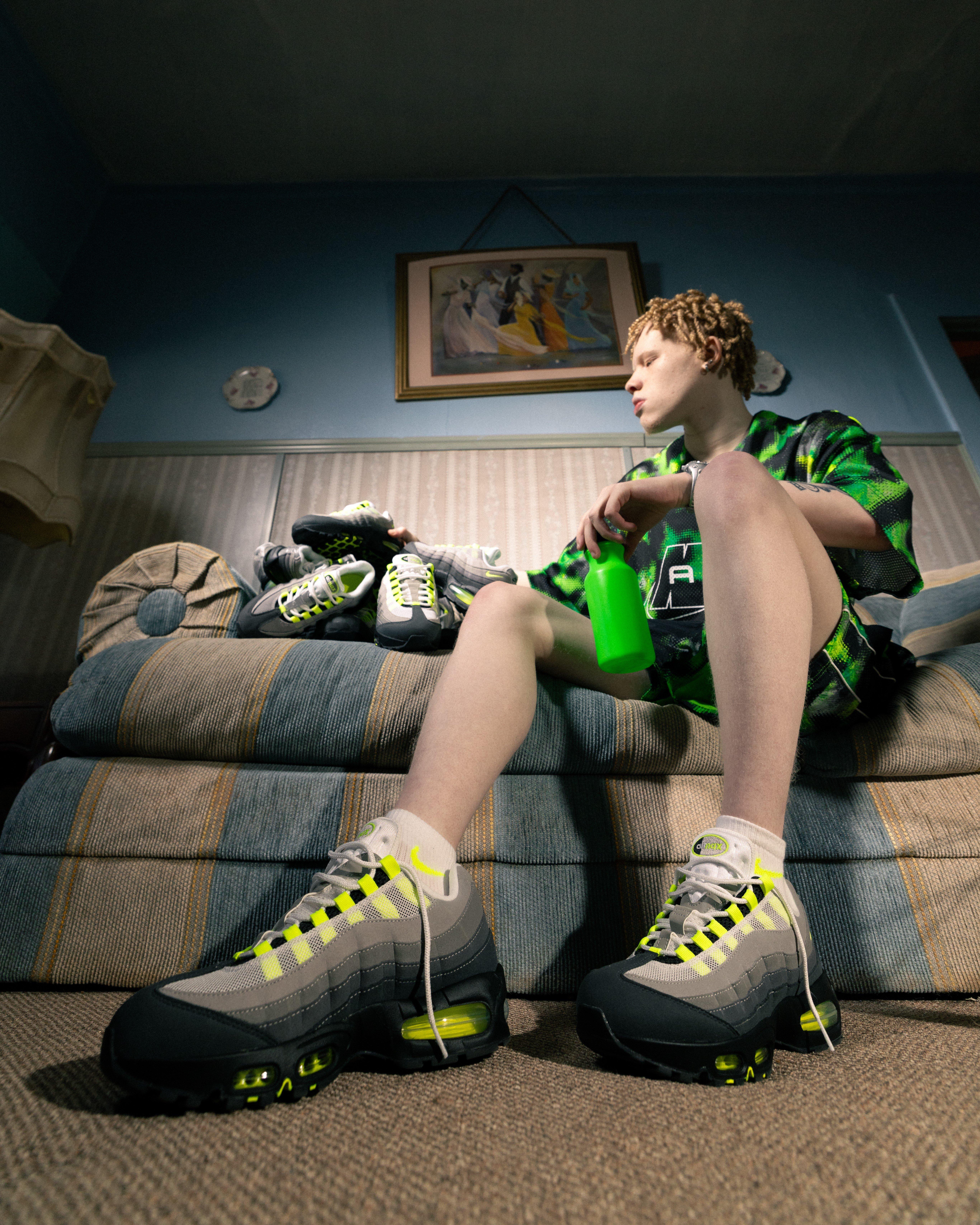 A low-angle shot of the same young man sitting on the edge of the striped sofa, holding a bright green water bottle. He is wearing the grey and neon yellow sneakers with white socks. The pile of sneakers remains on the sofa behind him, and a framed picture hangs on the blue wall above.