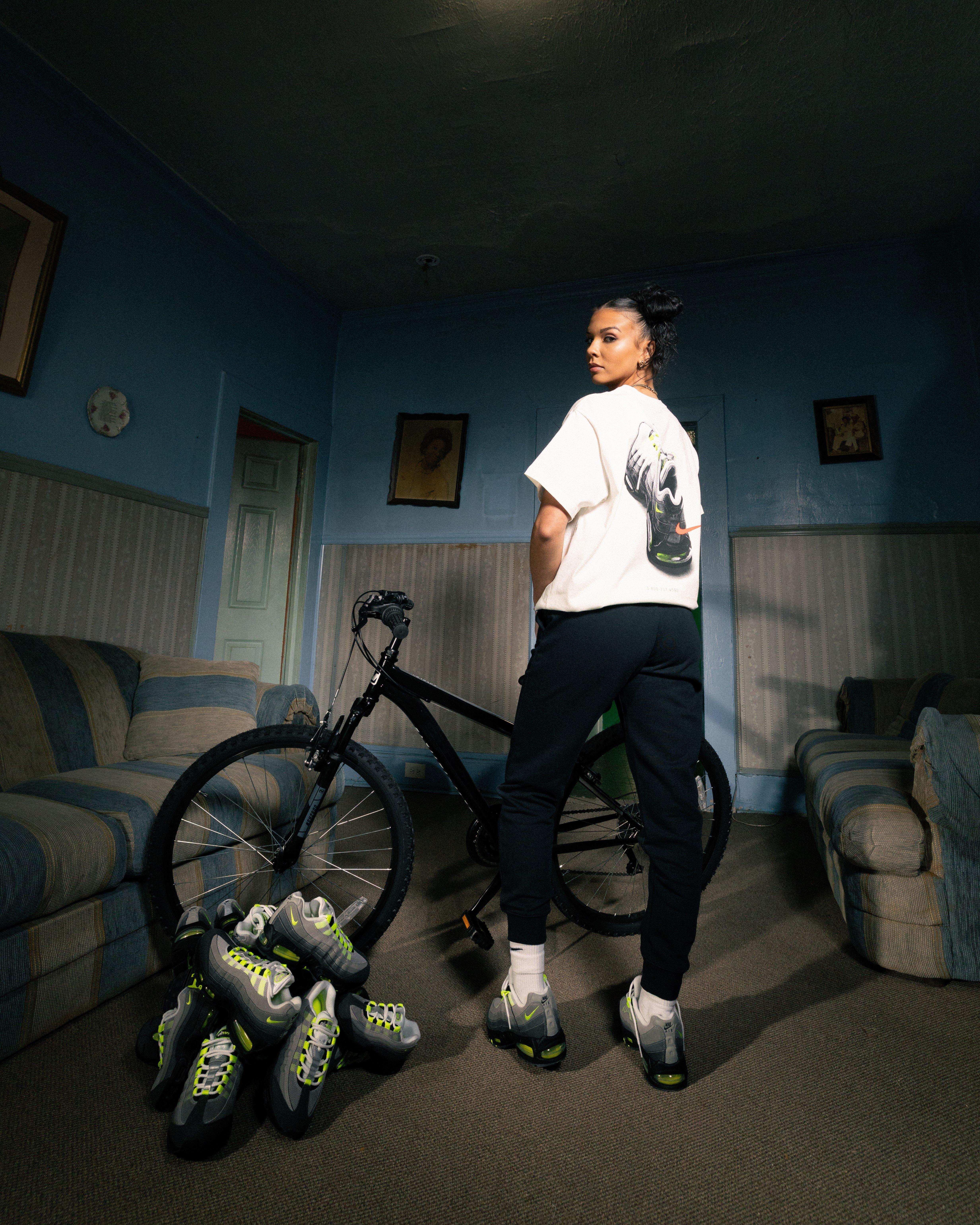 A young woman with her hair in two buns stands with her back to the camera, looking over her shoulder. She is wearing a white t-shirt featuring a large graphic of a Nike Air Max 95 on the back, paired with black jogger pants and the same grey and neon yellow sneakers. A black mountain bike and a pile of sneakers are on the floor next to her in a room with blue walls.