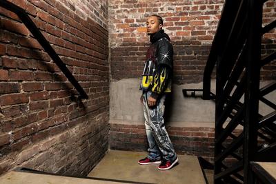 Man wearing a graphic black hoodie, distressed jeans, and red sneakers, standing in a brick stairwell with industrial details.