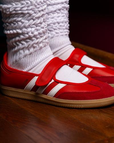 Close-up of red adidas Samba Mary Jane sneakers with white three stripes and gum sole, styled with white tights and chunky knit leg warmers on a wooden floor.