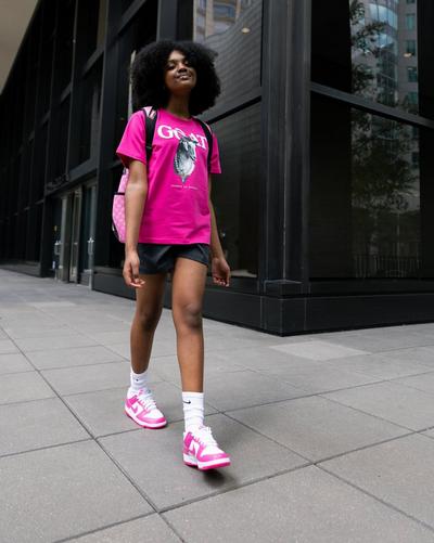 oung girl walking confidently in an urban setting, wearing a bright pink GOAT graphic t-shirt, black shorts, white Nike socks, and white and pink Nike sneakers with a pink backpack.