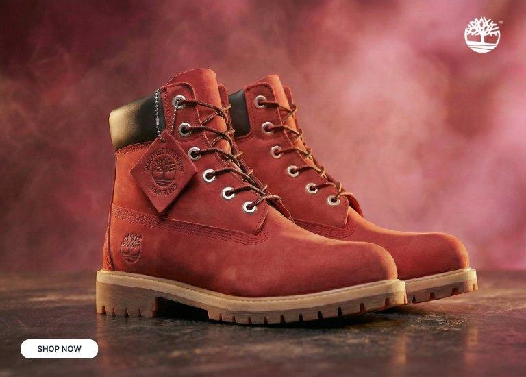 A pair of red Timberland 6-inch premium waterproof boots with black padded collars and silver eyelets, set against a smoky reddish-pink background with a "Shop Now" button in the corner.