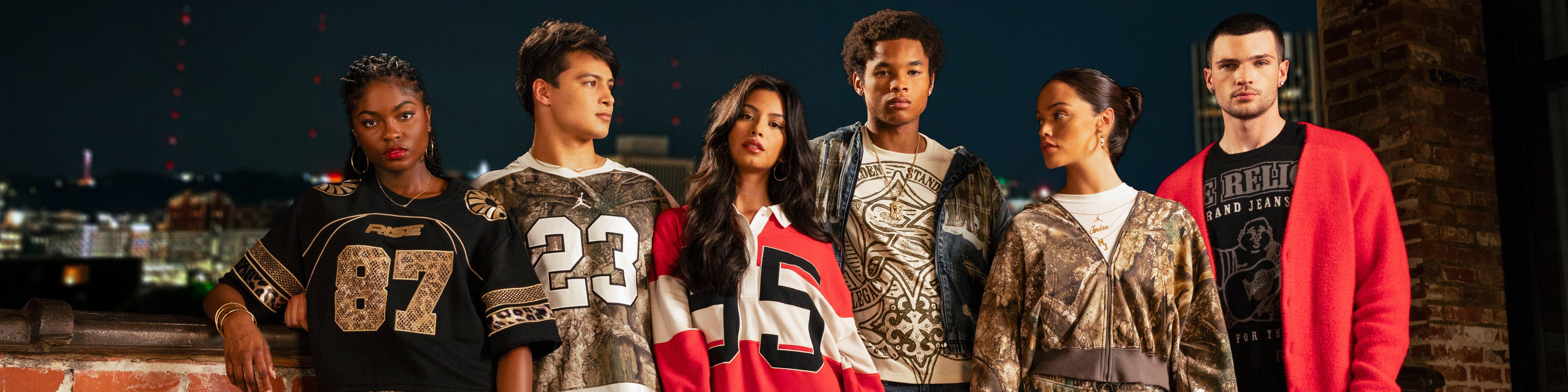 Group of six young adults standing together on a rooftop at night, wearing trendy streetwear and athletic-inspired outfits with bold prints, numbers, and camo patterns, city lights visible in the background.
