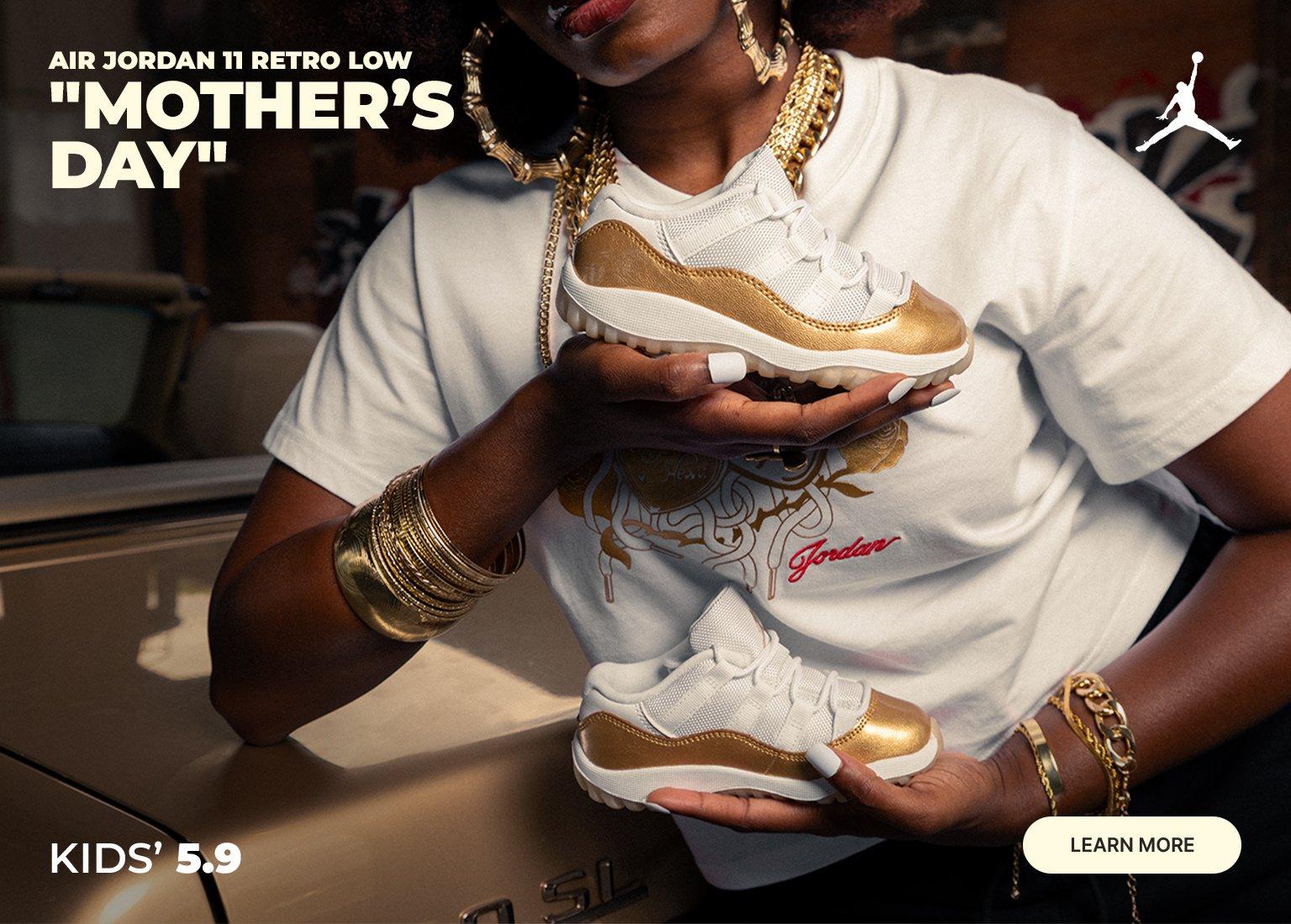  An adult holds a pair of kids' Air Jordan 11 Retro Low "Mother's Day" sneakers, which feature white mesh uppers and metallic gold mudguards. To the right, another pair of the gold and white shoes hangs by the laces. Text reads "KIDS' 5.9," "AIR JORDAN 11 RETRO LOW 'MOTHER'S DAY'," and a "LEARN MORE" button sits in the bottom corner.