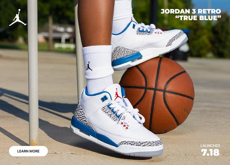 Jordan 3 Retro “True Blue” kids’ sneakers in white and blue displayed on an outdoor basketball court with a basketball. Graphic includes Jordan branding, a Learn More button, and launch date of 7/18.
