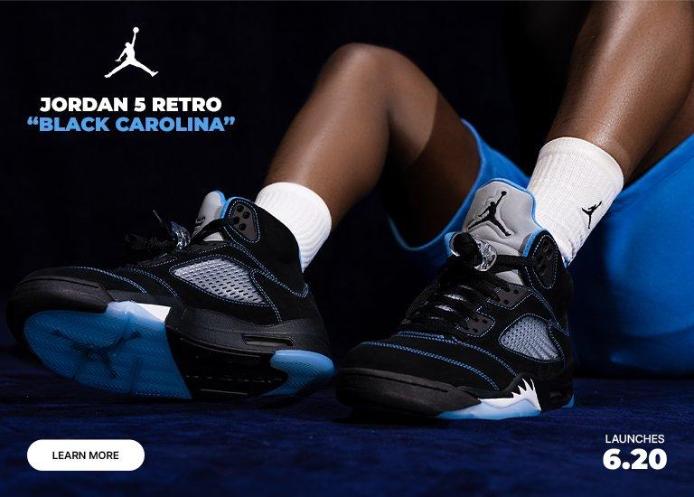 A close-up of a person sitting down wearing Jordan 5 Retro Black Carolina sneakers, white socks with the Jumpman logo, and blue shorts. Text highlights Jordan 5 Retro Black Carolina, Launches 6.20, and includes a Learn More button.