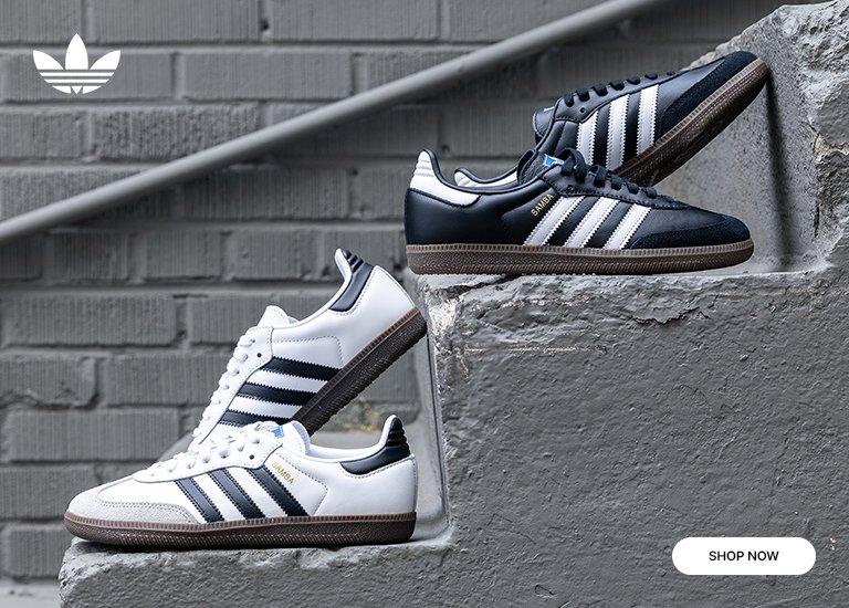 Three pairs of Adidas Samba sneakers are stacked on concrete steps against a gray brick wall. The top pair is black with white stripes, the middle pair is white with black stripes and a dark sole, and the bottom pair is white with black detailing. The Adidas trefoil logo appears in the top left corner, and a “Shop Now” button is in the bottom right corner.