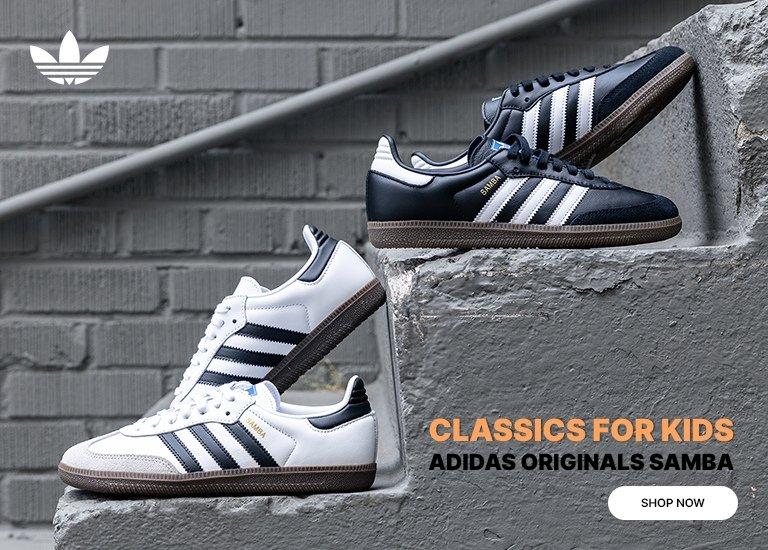  An advertisement for kids' Adidas Originals Samba sneakers featuring two pairs—one black with white stripes and one white with black stripes—posed on concrete stairs against a brick wall. The image includes the Adidas logo, the text "Classics for Kids," and a "Shop Now" button.