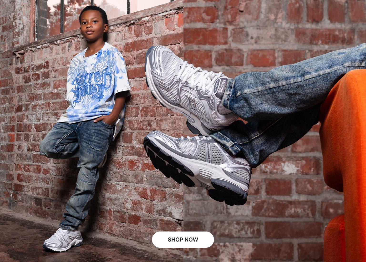 Kids' graphic tee with light-wash jeans and silver Saucony sneakers in an industrial brick loft setting.