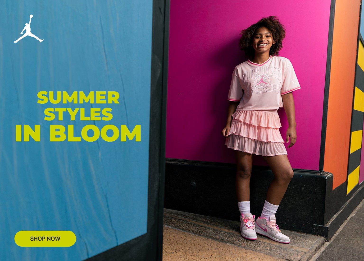 A three-panel promotional banner features a young girl smiling and posing in a pink Jordan crop top and a layered ruffle skirt. The left panel shows her from the waist up against a bright green background with a white Jordan Jumpman logo in the top corner. The middle panel features a close-up of an Air Jordan 1 Mid sneaker in pink and grey texture with a bright pink Swoosh and colorful lace charms against a magenta background with yellow text reading SUMMER STYLES IN BLOOM. The right panel shows her full-body pose against a magenta and multicolored wall, with a lime green SHOP NOW button in the bottom corner.
