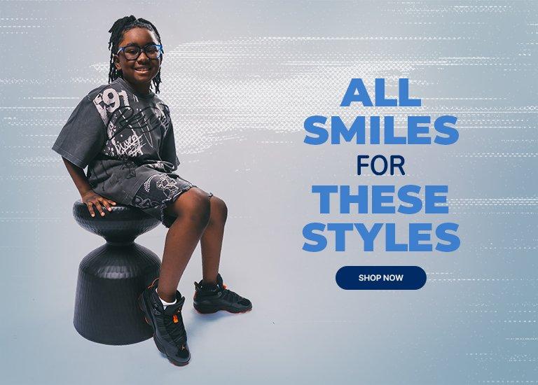 Smiling child wearing a black graphic outfit and black sneakers sits on a stool beside text reading “All Smiles for These Styles” with a “Shop Now” button.