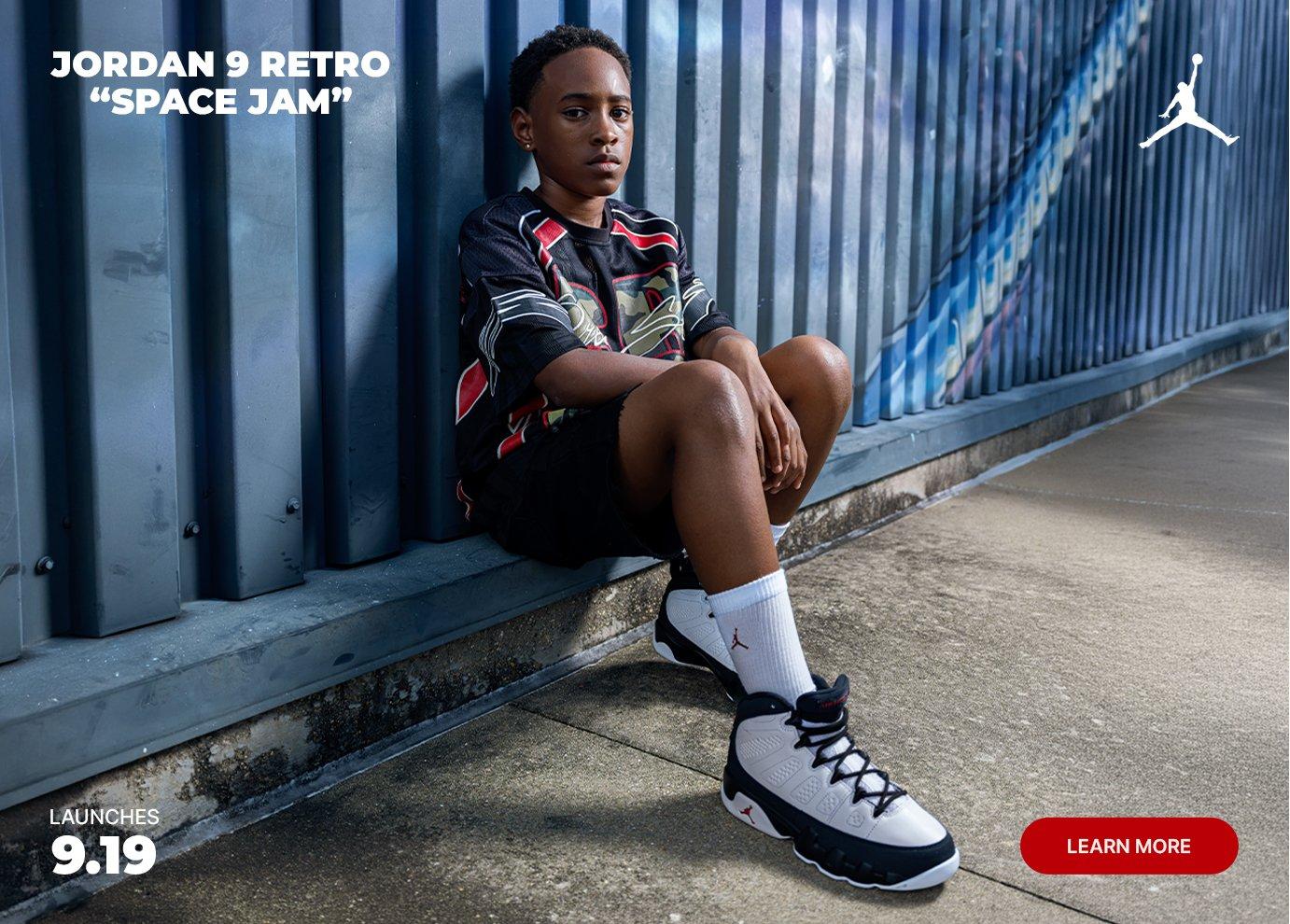 Kid wearing Jordan 9 Retro White/Black sneakers sits against a blue wall in a lifestyle campaign image.