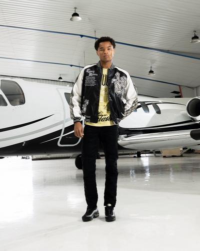 Traveler wearing a black-and-white graphic jacket, black pants and sneakers while standing beside a white private plane in a hangar.