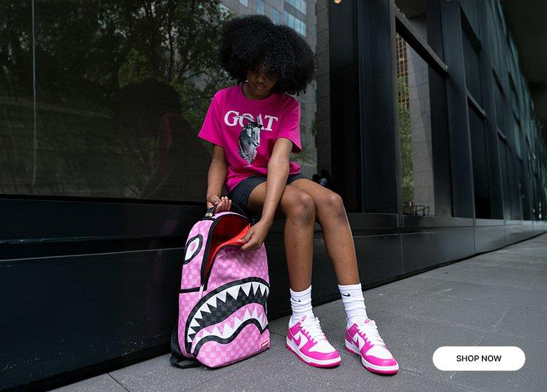 A girl is outdoors, wearing Streetz Iz Watchin GOAT Big Girls' Tee, black shorts, white Nike socks, and Nike Dunk Low "White/Laser Fuchsia" Shoe. She has a Sprayground Shark Pink Drip Backpack. "SHOP NOW" 