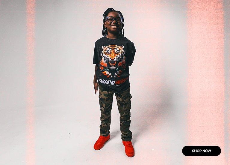A young person smiling in streetwear, featuring a black Show No Mercy tiger graphic tee, camo cargo pants, and bright red sneakers against a studio backdrop with a Shop Now button.