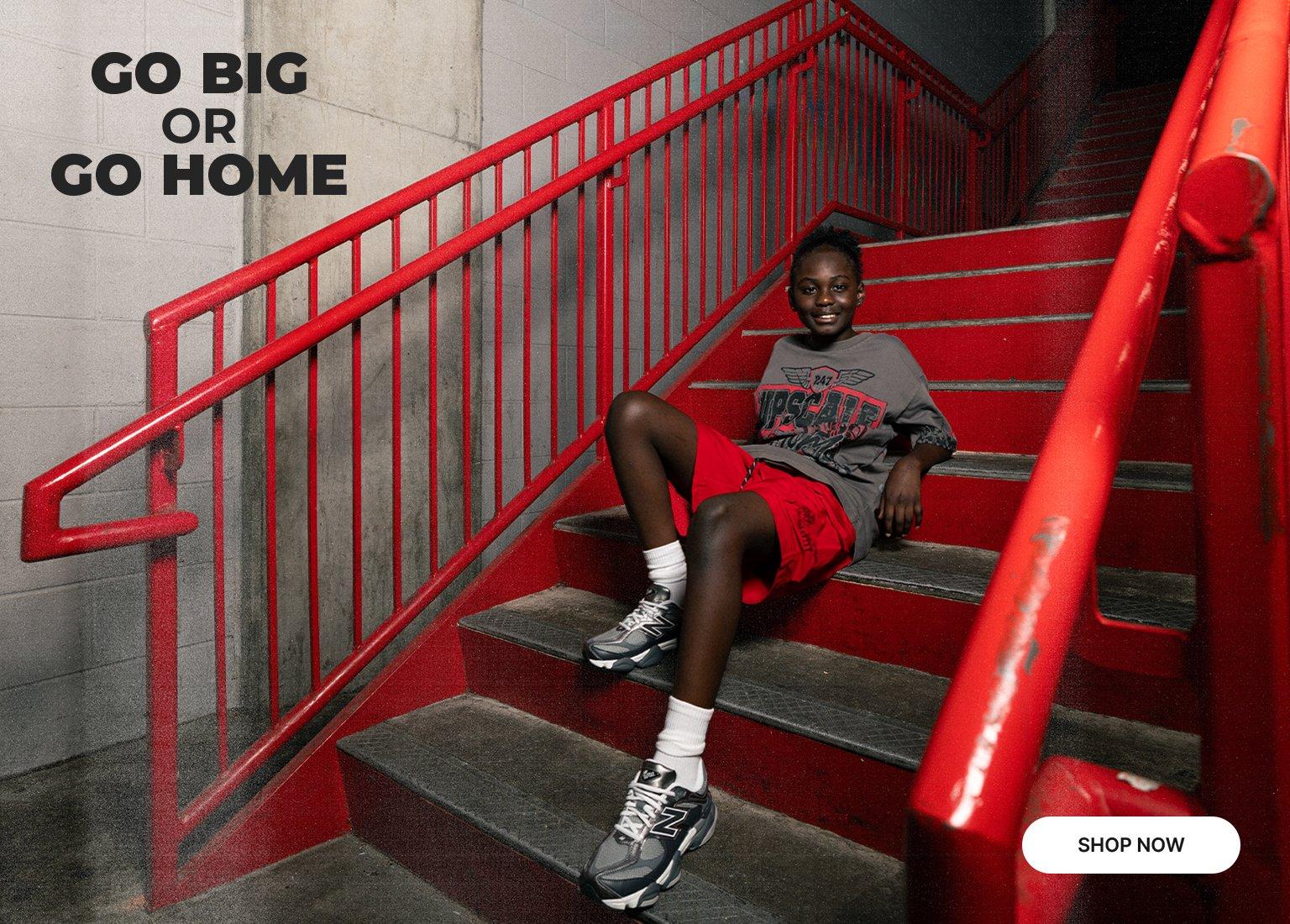 A split banner featuring a youth posing on red stairs wearing a grey graphic t-shirt, red shorts, and grey New Balance sneakers. Text on the left reads, GO BIG OR GO HOME, with a SHOP NOW button.