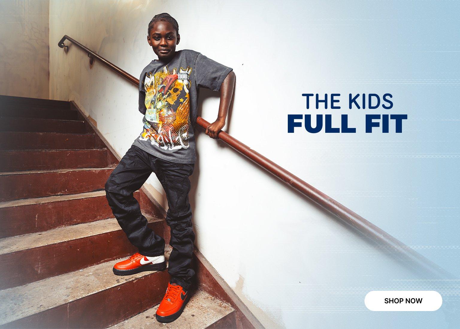 Promotional banner featuring a child modeling a graphic t-shirt and black pants while leaning against a handrail on a staircase. The child is wearing orange and black Nike Air Force 1 Low sneakers. To the right, the text reads THE KIDS FULL FIT in navy blue, and a Shop Now button is located in the bottom right corner.