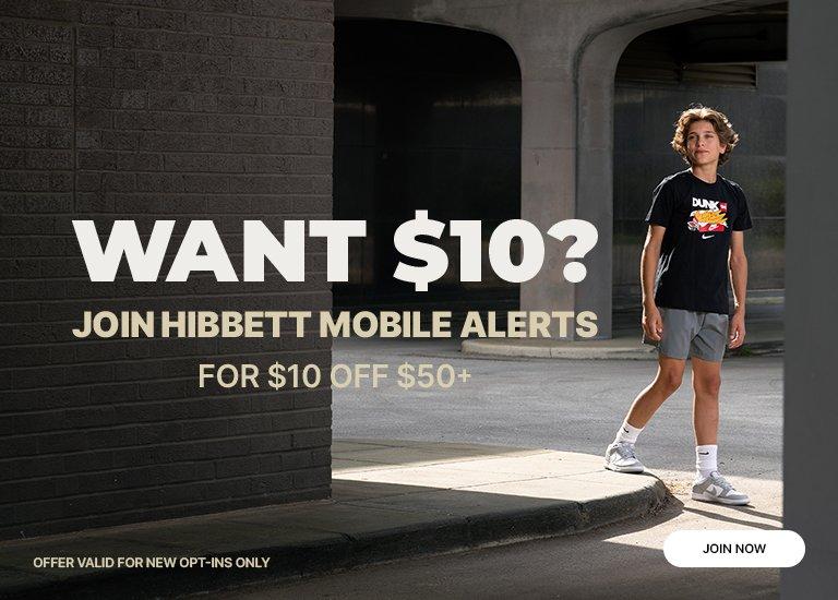 A boy in a black T-shirt and gray shorts stands on a curb, looking at the camera. He wears a pair of white sneakers. Behind him is a building with the text, WANT $10? JOIN HIBBETT MOBILE ALERTS FOR $10 OFF $50+. The text OFFER VALID FOR NEW OPT-INS ONLY is below.