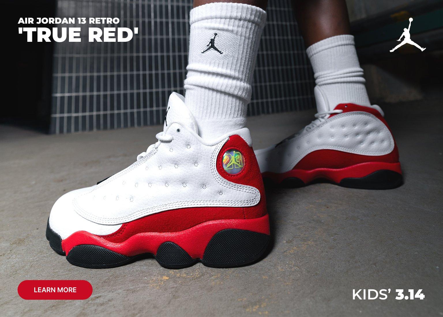 A split-screen promotional banner featuring the Air Jordan 13 Retro True Red. The left side shows a close-up of the white and red sneakers being worn with white crew socks. The right side shows a young boy standing in a doorway wearing the sneakers, red shorts, and a graphic t-shirt. Text includes Air Jordan 13 Retro True Red, Kids 3.14, and a Learn More button.