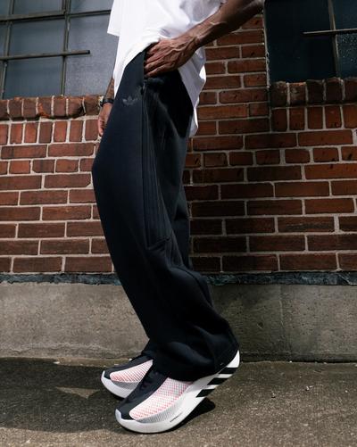 Person wearing black adidas track pants and white, black and red adidas Hyperboost Edge running shoes beside a brick wall.