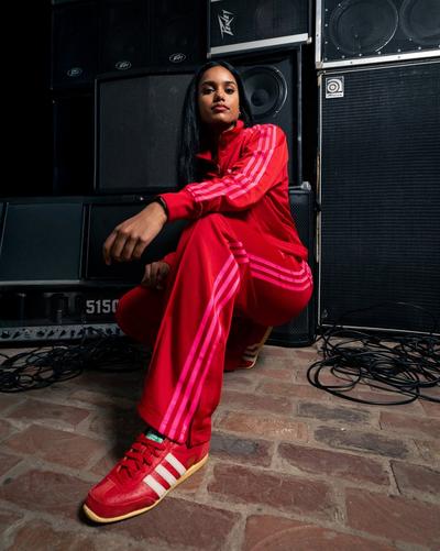 A woman with long dark hair is crouching down in front of a stack of large black speakers and amplifiers. She is wearing a red Adidas tracksuit with pink stripes and matching red sneakers with white laces.