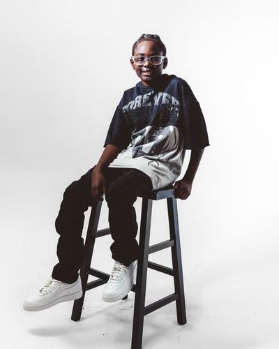 Kid sitting on a stool in a graphic oversized T-shirt, black pants, and white Nike Air Force 1 sneakers.  Provide your feedback on BizChat