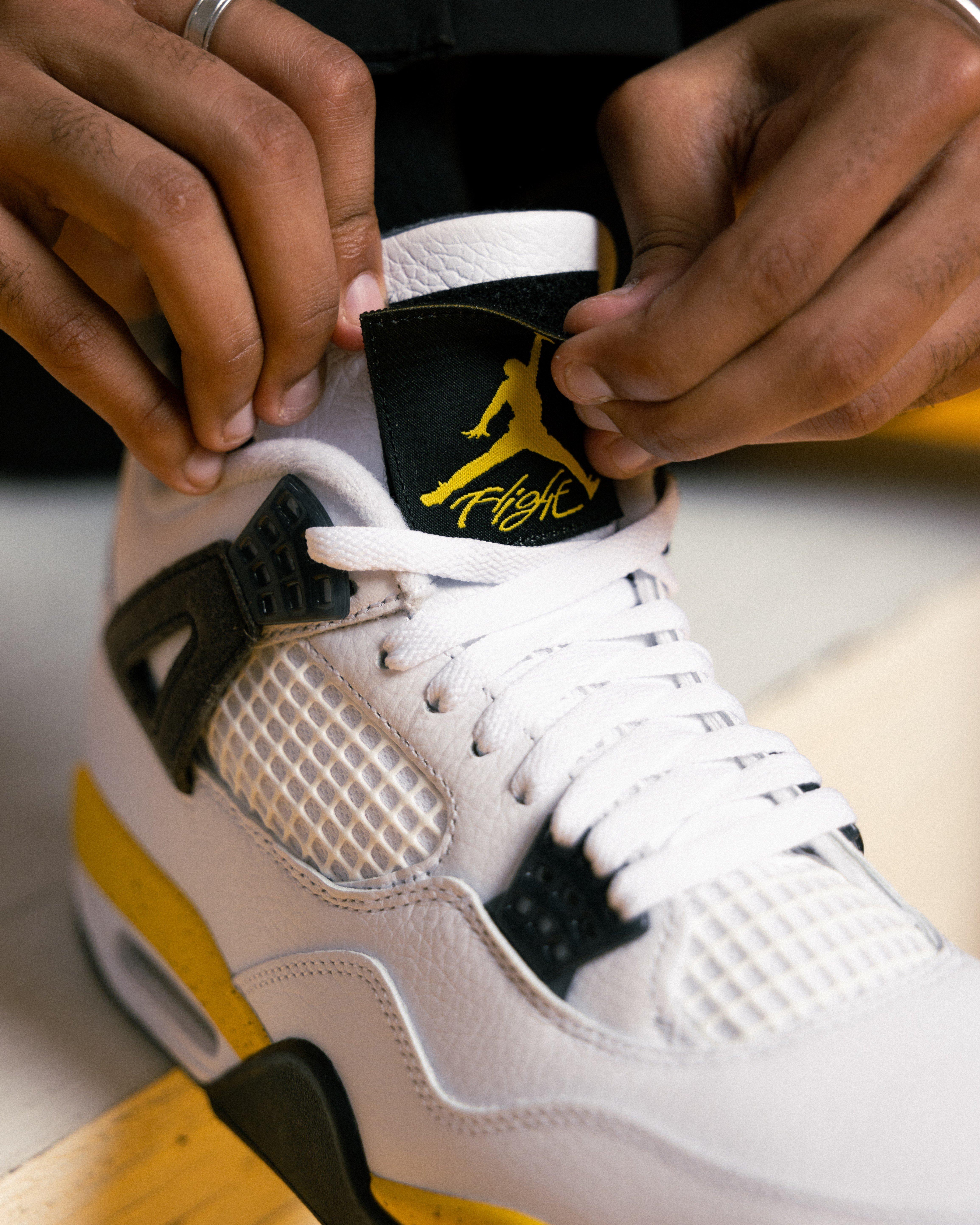Close-up of an Air Jordan 4 Tour Yellow sneaker showing white laces, white mesh, black lace wings and a removable black tongue patch with a yellow Jumpman Flight logo.