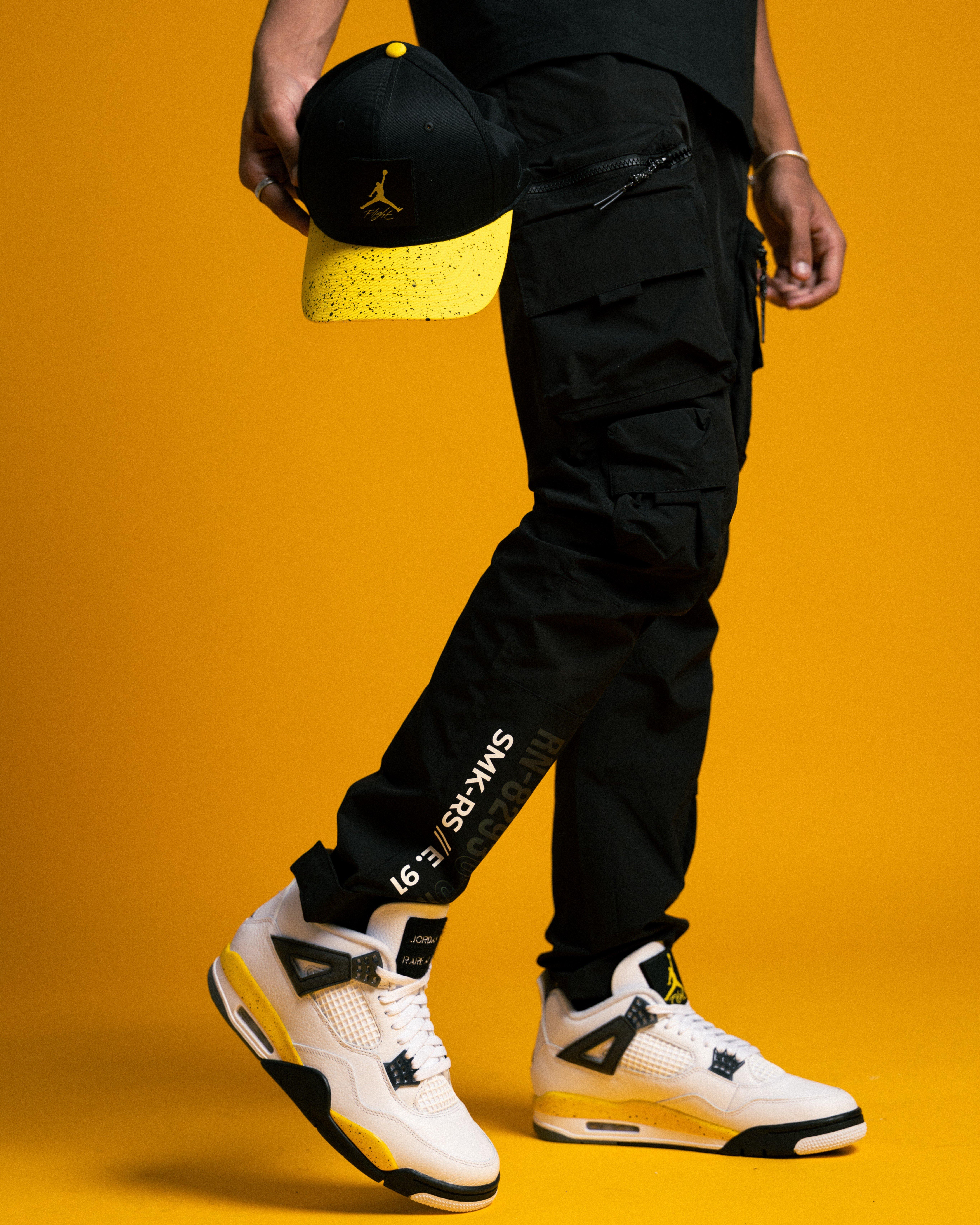 Person wearing Air Jordan 4 Tour Yellow sneakers with black cargo pants while holding a black-and-yellow Jordan Flight cap against a yellow background.