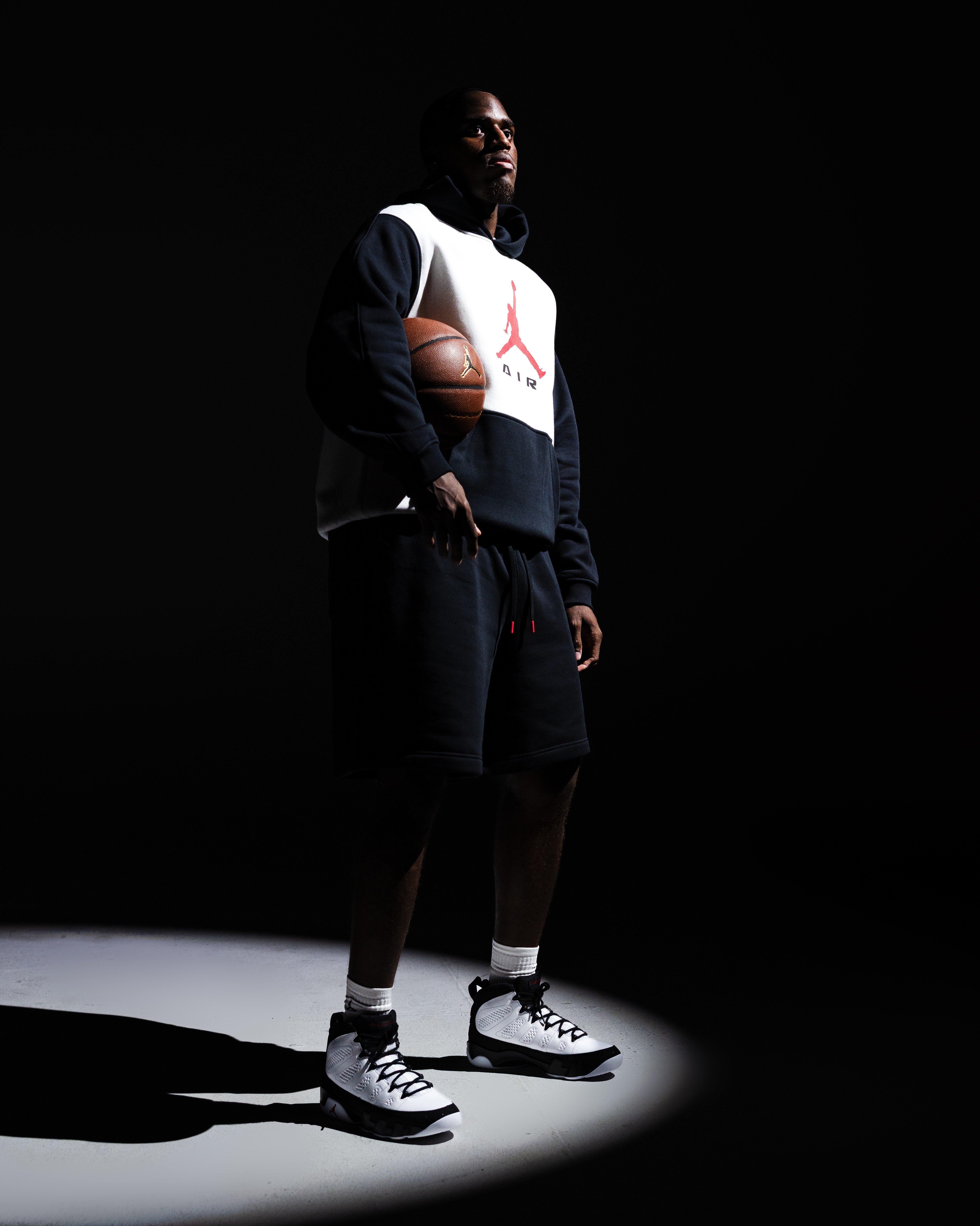 Person standing under a spotlight holding a basketball and wearing a navy-and-white Jordan hoodie, navy shorts and black-and-white Air Jordan 9 Retro Space Jam sneakers.