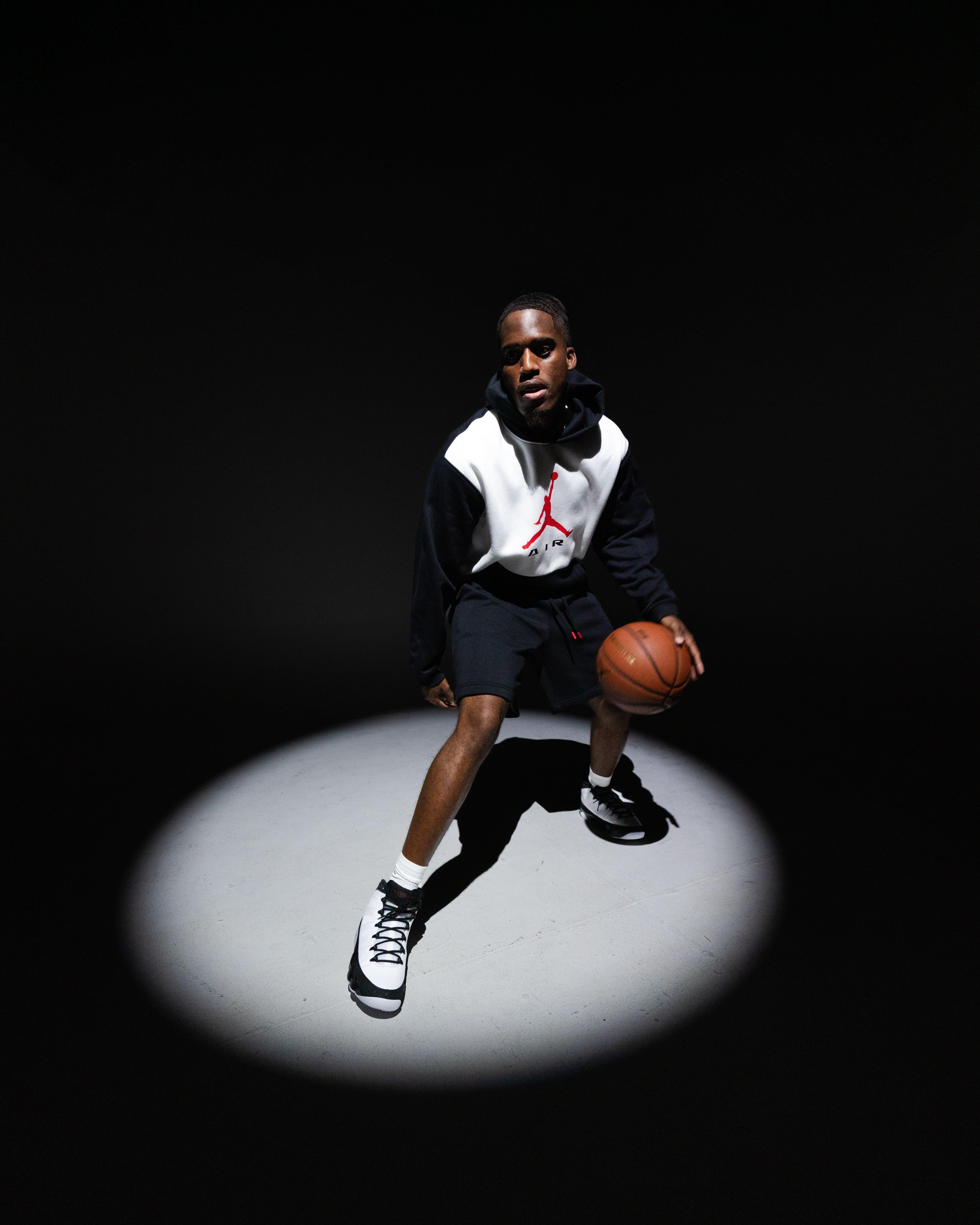 Person dribbling a basketball under a spotlight while wearing a navy-and-white Jordan hoodie, navy shorts and Air Jordan 9 Retro Space Jam sneakers.