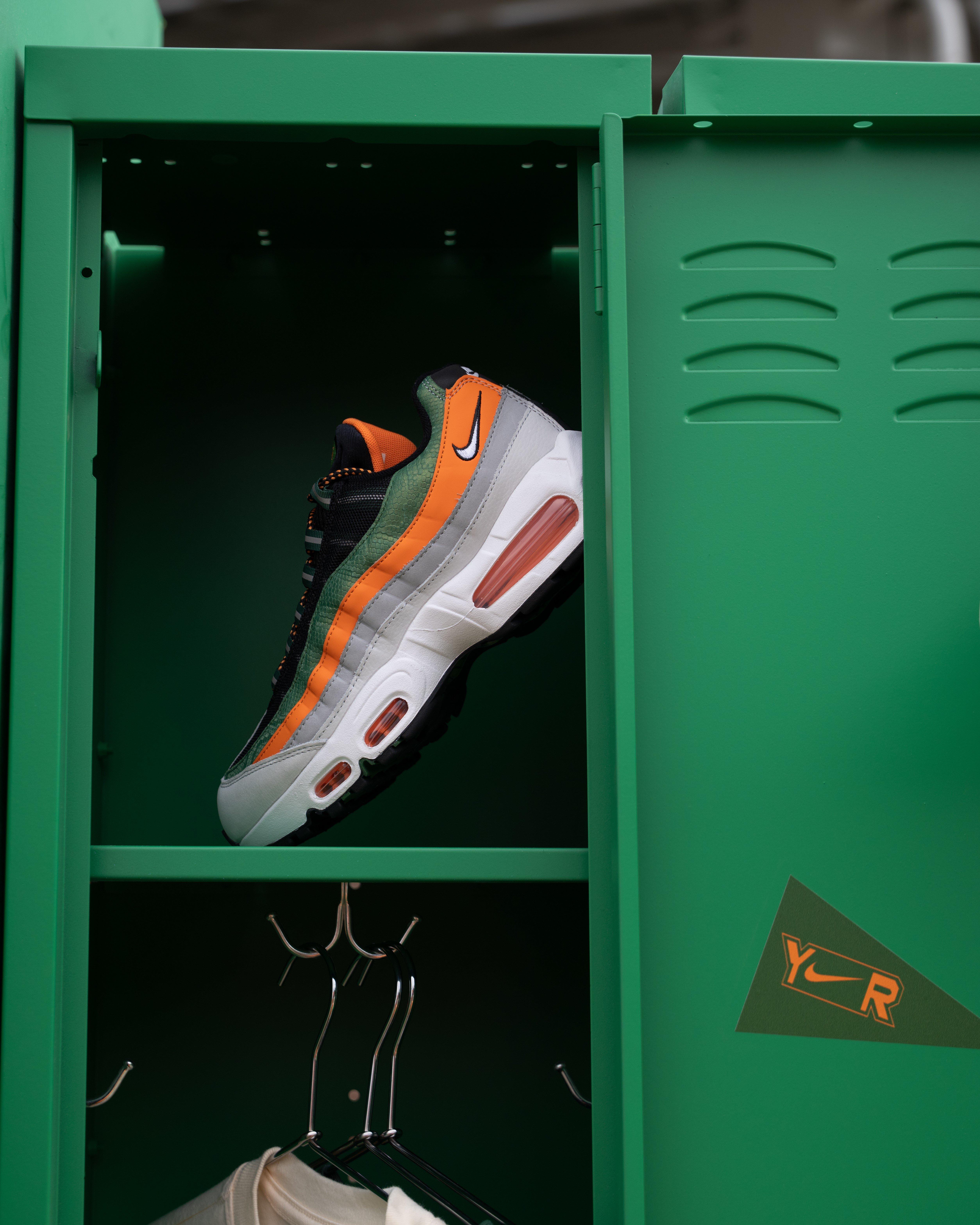 A close-up of a single Nike Air Max 95 sneaker in orange, green, black, and white, resting on a shelf inside a green metal locker with a Y-R logo.