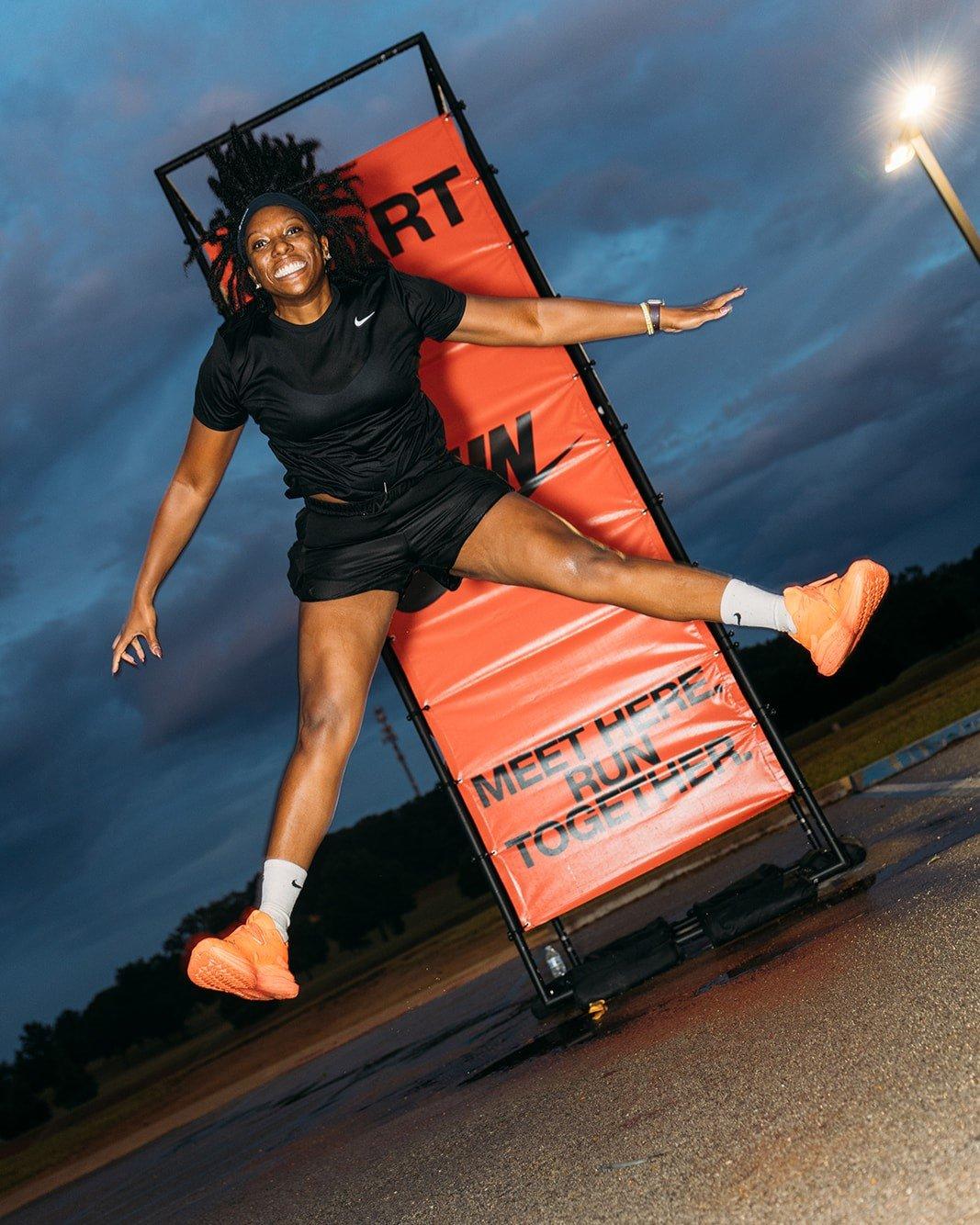 A woman with dreadlocks, wearing all black athletic wear, is mid-air, jumping in front of a large orange banner that says "START RUN". She is smiling and has her arms and legs extended out in a star shape. The sky behind her is dark and cloudy.
