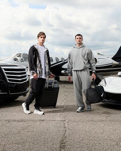 Two people pose on an airport tarmac with luggage, wearing layered Nike travel outfits and sneakers beside parked vehicles and private planes.